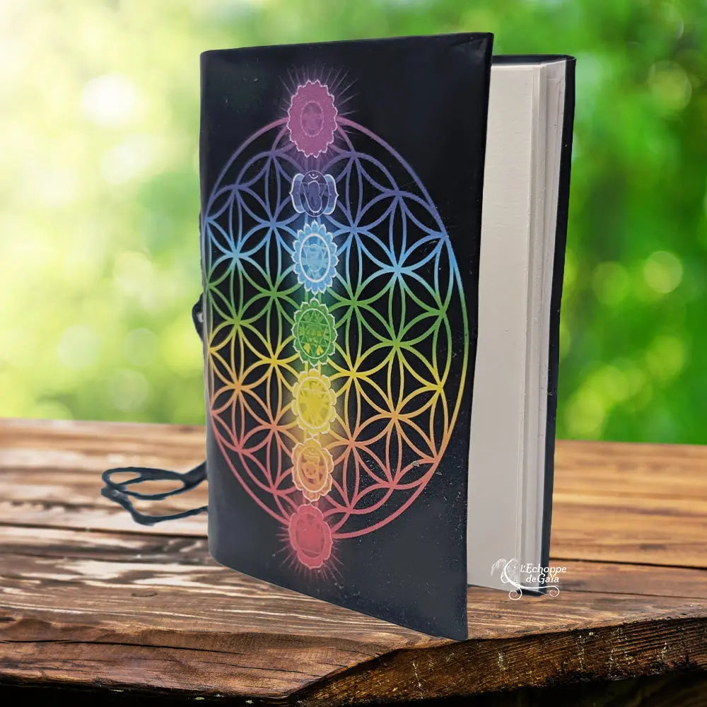 Un carnet de notes à couverture noire illustrée d'une fleur de vie arc-en-ciel et des symboles des sept chakras, muni d'un cordon de fermeture et posé sur un support en bois.