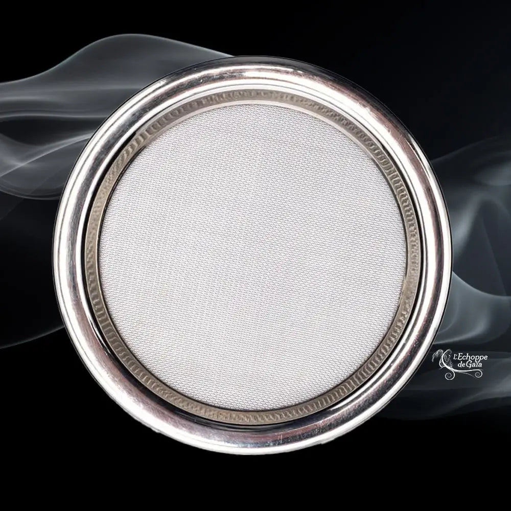 Grand tamis rond en métal argenté avec grille fine, photographié sur fond noir avec volutes de fumée.