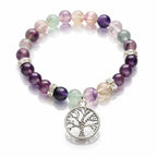 Un bracelet en pierres naturelles de fluorite, alternant des perles rondes multicolores (violet, vert, translucide), orné d'un pendentif Arbre de Vie en métal argenté et de perles de strass, présenté sur un fond blanc réfléchissant.