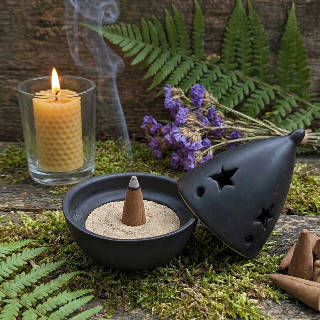 Brûle-encens en céramique noire avec couvercle ajouré d’étoiles, cône sur sable et fumée visible, bougie en verre, fougère et fleurs violettes.