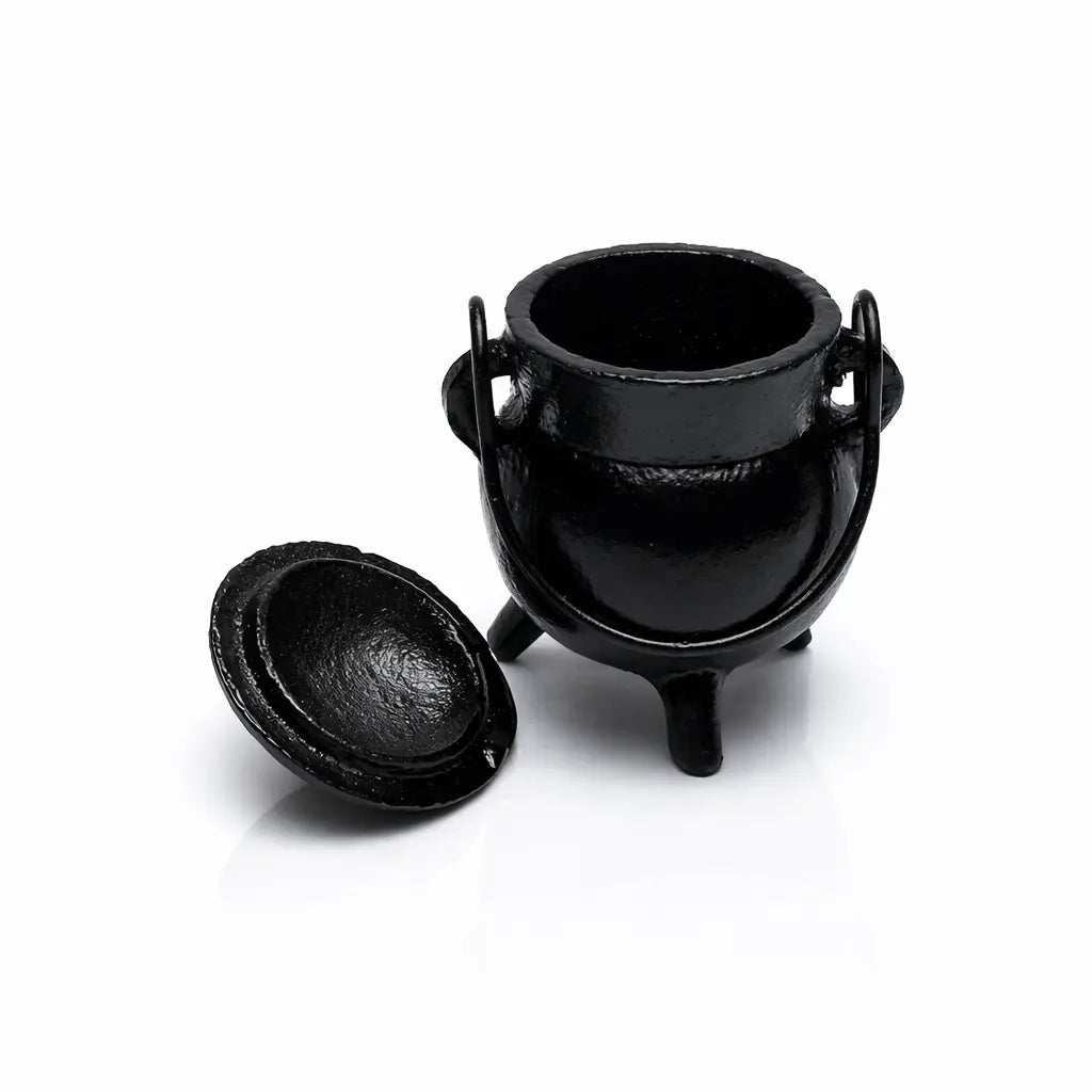 Un mini chaudron de sorcière en fonte noire, doté d'une anse et de trois pieds, avec son couvercle posé à côté, présenté en vue plongeante sur un fond blanc réfléchissant.