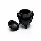 Un mini chaudron de sorcière en fonte noire, doté d'une anse et de trois pieds, avec son couvercle posé à côté, présenté en vue plongeante sur un fond blanc réfléchissant.