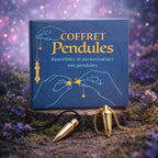 Boîte de coffret bleue "Coffret Pendules" posée dans la nature, accompagnée de deux pendules dorés de formes différentes, l'un sur cordon noir et l'autre sur chaîne dorée.