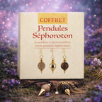 Boîte de coffret beige "Coffret Pendules Séphoroton" posée dans la nature, accompagnée de deux pendules Séphoroton, l'un en bois sur cordon noir et l'autre en œil de tigre sur chaîne dorée.