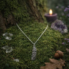 Collier argenté avec un pendentif en forme de plume finement ciselée, déposé sur de la mousse forestière à côté d'une bougie violette, de cristaux et d'un gland.