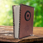 Un carnet grimoire en cuir marron entrouvert, bordé d'un tressage en cuir noir et orné d'une rose des vents ainsi que d'une pierre d'onyx centrale, présenté sur un support en bois.