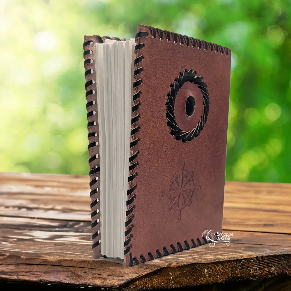 Un carnet grimoire en cuir marron entrouvert, bordé d'un tressage en cuir noir et orné d'une rose des vents ainsi que d'une pierre d'onyx centrale, présenté sur un support en bois.