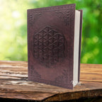 Un carnet grimoire en cuir marron foncé entrouvert, dont la couverture est ornée d'une fleur de vie et de motifs d'angles embossés, présenté sur un support en bois.