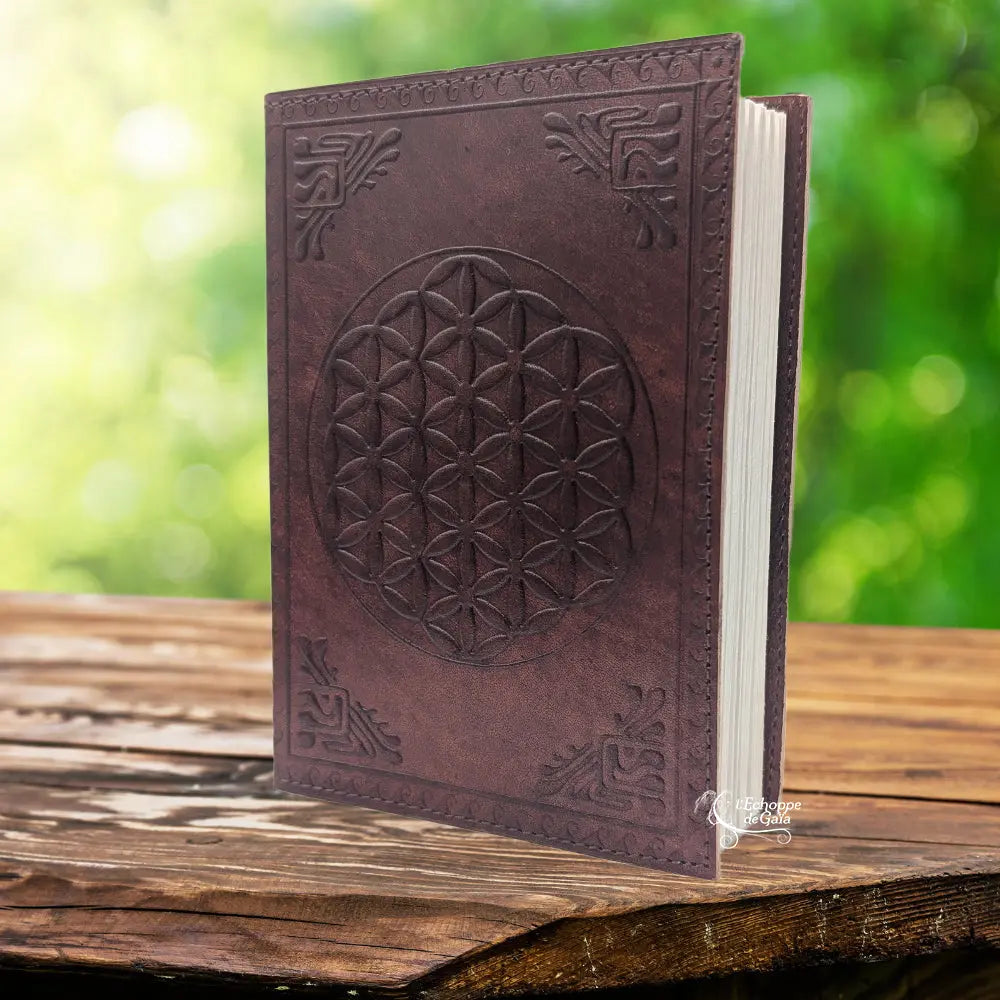 Un carnet grimoire en cuir marron foncé entrouvert, dont la couverture est ornée d'une fleur de vie et de motifs d'angles embossés, présenté sur un support en bois.