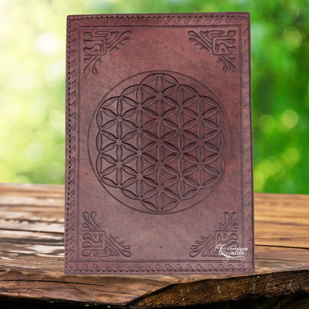 La couverture d'un carnet grimoire en cuir marron foncé ornée d'une fleur de vie centrale et de motifs d'angles finement embossés, présentée sur un support en bois.