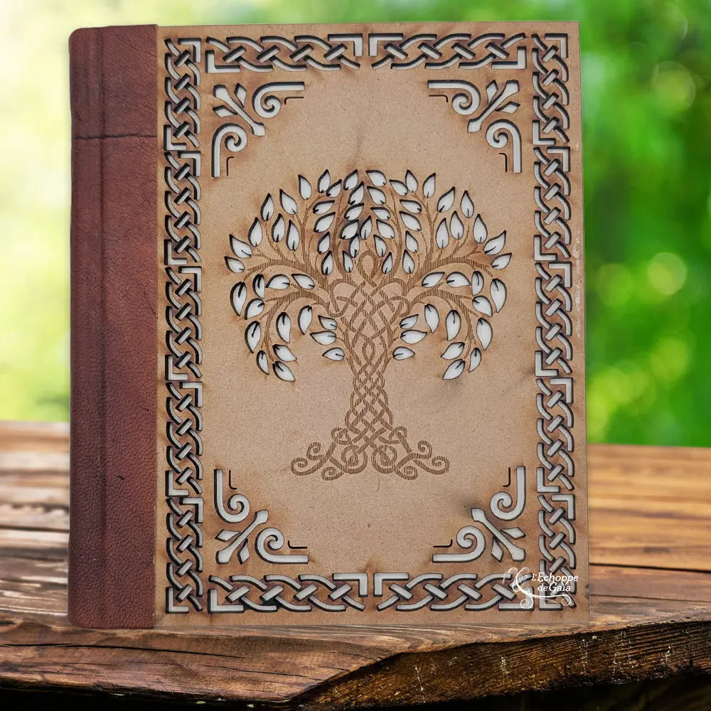 La couverture d'un carnet grimoire associant un dos en cuir marron et une plaque de bois finement sculptée d'un arbre de vie central et de bordures celtiques, présentée sur un support en bois.