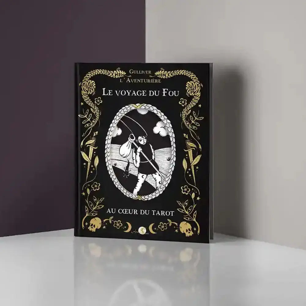 Livre Le Voyage du Fou posé debout, couverture noire avec ornements dorés et illustration du Fou, sur fond gris.