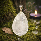 Pendentif ovale en cristal de roche translucide avec de fines aiguilles de tourmaline noire à l'intérieur, posé sur de la mousse avec une bougie allumée.