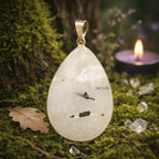 Un pendentif en cristal de roche en forme de goutte, contenant des inclusions de tourmaline noire, surmonté d'une bélière en métal argenté et mis en scène dans un décor naturel de mousse avec une bougie allumée.
