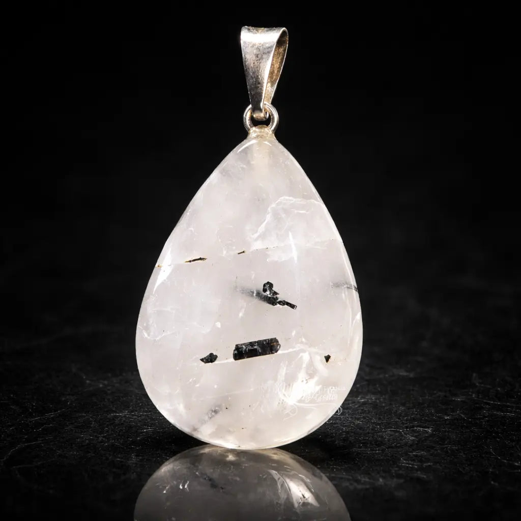 Un pendentif en cristal de roche en forme de goutte, contenant des inclusions de tourmaline noire, surmonté d'une bélière en métal argenté et présenté avec son reflet sur un fond noir.