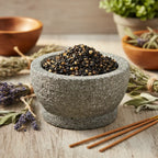 Grains d'encens de résine Pontifical (noir et ambre) dans un mortier en granit gris, entouré de bouquets de lavande, de feuilles de sauge et de bâtonnets d'encens.