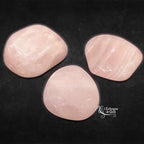 Trois pierres de quartz rose roulées aux formes généreuses et aux teintes rose pâle délicates, disposées sur un fond noir texturé.