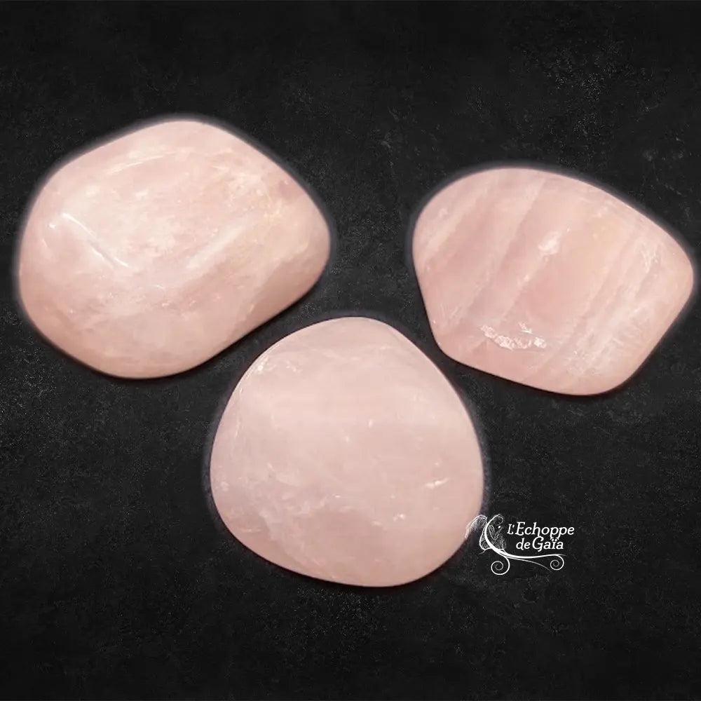 Trois pierres de quartz rose roulées aux formes généreuses et aux teintes rose pâle délicates, disposées sur un fond noir texturé.