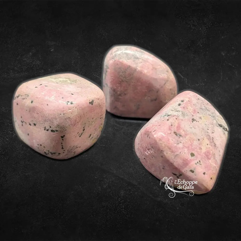Trois pierres de rhodonite polies, affichant des teintes vieux rose avec des marbrures grises, noires et blanchâtres, présentées sur un fond noir.
