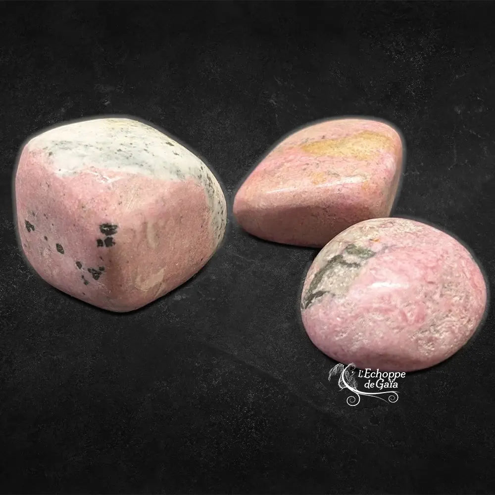 Trois pierres de rhodonite polies, affichant des teintes vieux rose avec des marbrures grises, noires et blanchâtres, présentées sur un fond noir.