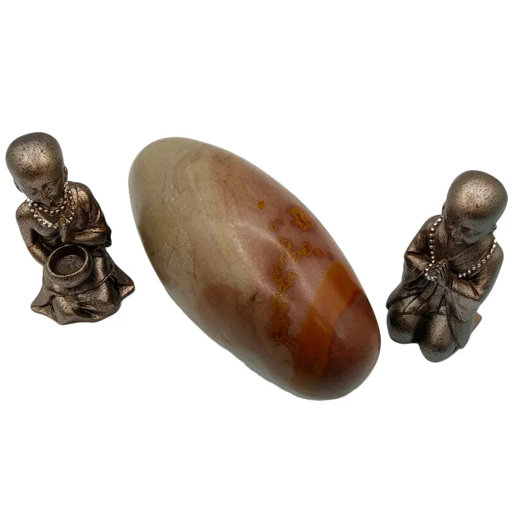 Pierre de Shiva Lingam polie de forme ovoïde, aux nuances naturelles beiges et brunes, disposée entre deux statuettes de moines bouddhistes en prière sur un fond blanc.