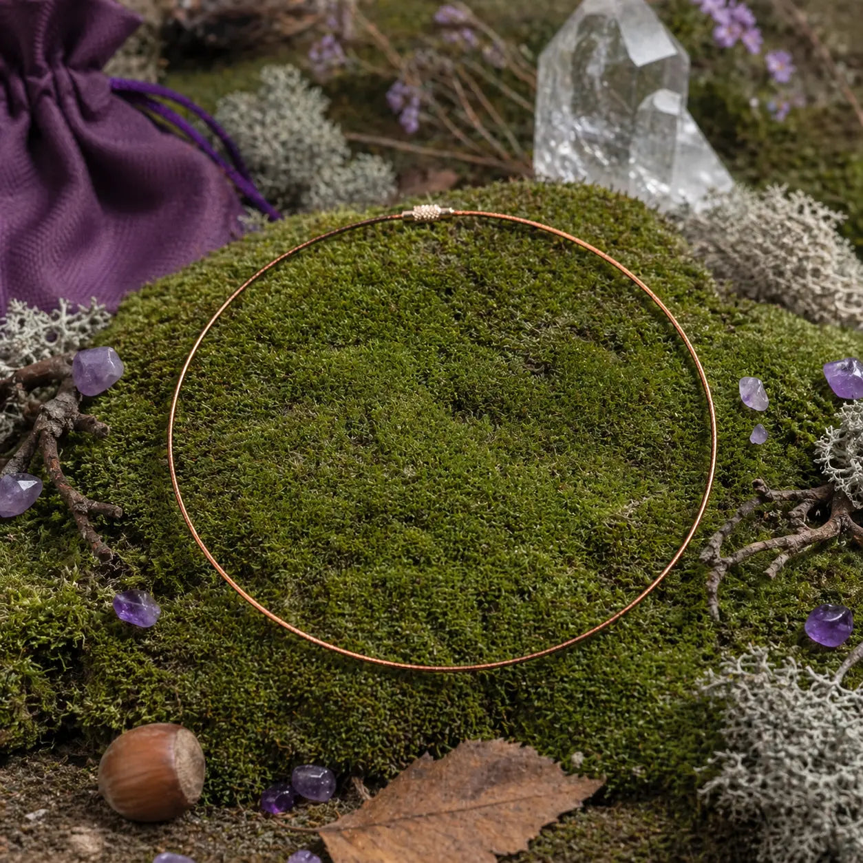 Câble gainé marron circulaire avec fermoir argenté, posé sur une souche de bois moussue entourée de fleurs violettes dans une ambiance de forêt sacrée.