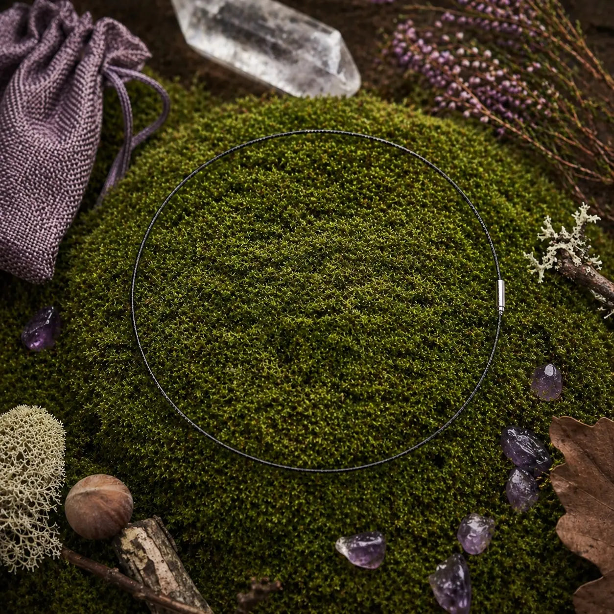 Câble gainé noir circulaire avec fermoir argenté, posé sur une butte de mousse avec une pointe de cristal, une pochette violette et des pierres d'améthyste.