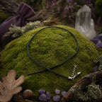 Cordon en cuir noir souple croisé sur une butte de mousse verdoyante, accompagné d'une pochette violette, d'une pointe de cristal et de petites améthystes.