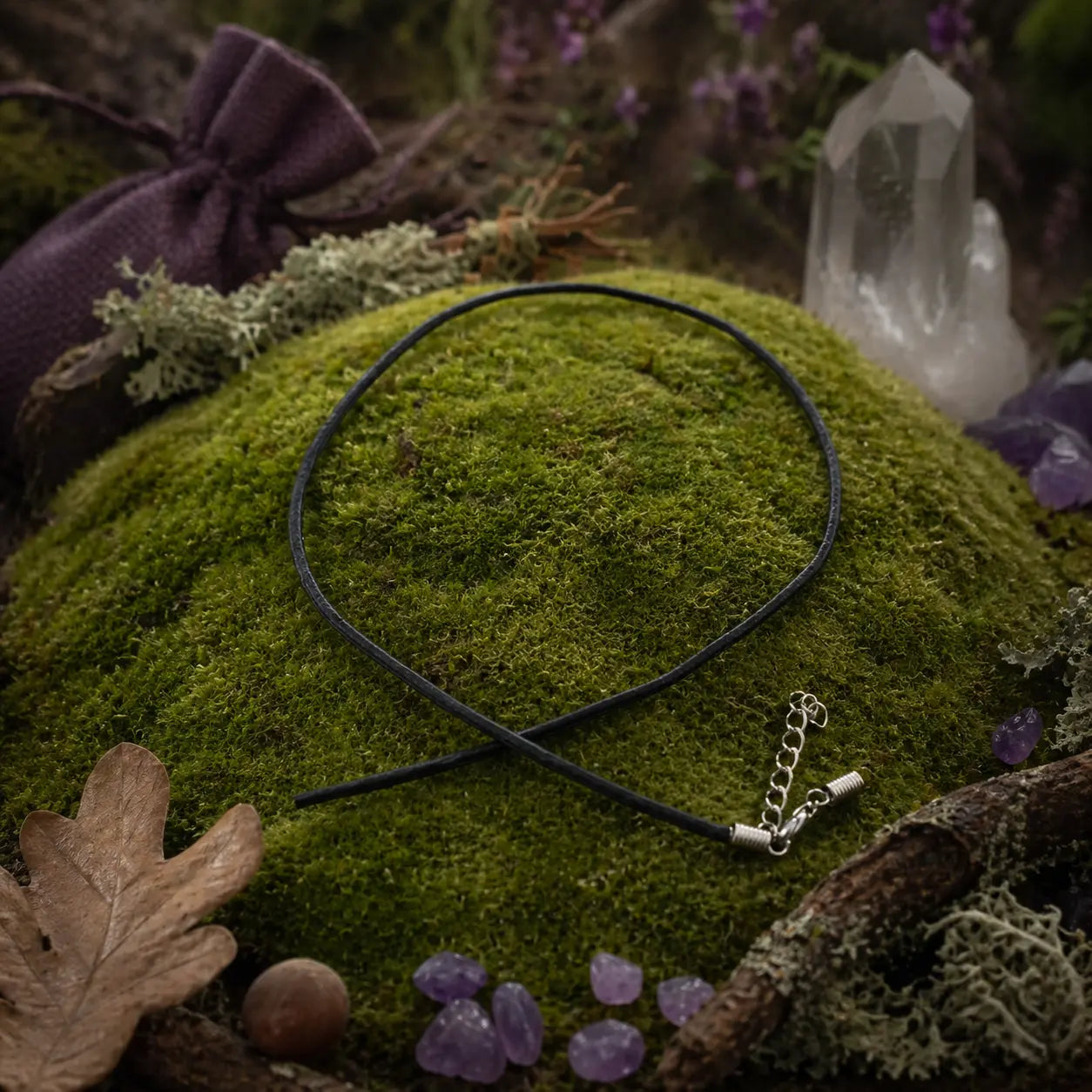 Cordon en cuir noir souple croisé sur une butte de mousse verdoyante, accompagné d'une pochette violette, d'une pointe de cristal et de petites améthystes.