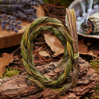 Tresse purificatrice de foin d’odeur (sweet grass) formant un petit cercle dressé sur un morceau d’écorce, brins noués sur le côté. En arrière-plan, feuilles mortes, aiguilles de pin et bâton de sauge fumant dans une coquille : ambiance rituelle de fumigation en forêt.