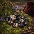 Sélection de pierres d'agate noire polies présentant des strates naturelles rubanées blanches et brunes, disposées sur de la mousse au pied d'un arbre avec un pochon bordeaux.