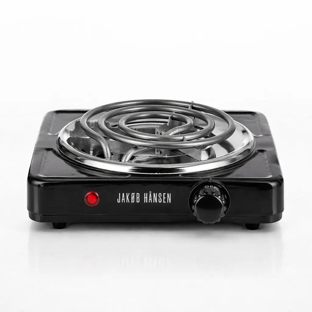 Un allume-charbon électrique noir de marque Jakob Hansen, muni d'une résistance en spirale, d'un bouton de réglage et d'un témoin lumineux rouge, présenté sur un fond blanc.