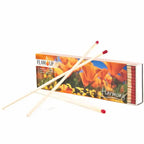 Boîte d'allumettes Flamup Harmony illustrée de fleurs orange, ouverte sur le côté et accompagnée de trois longues allumettes à tête rouge posées devant elle, sur fond blanc.
