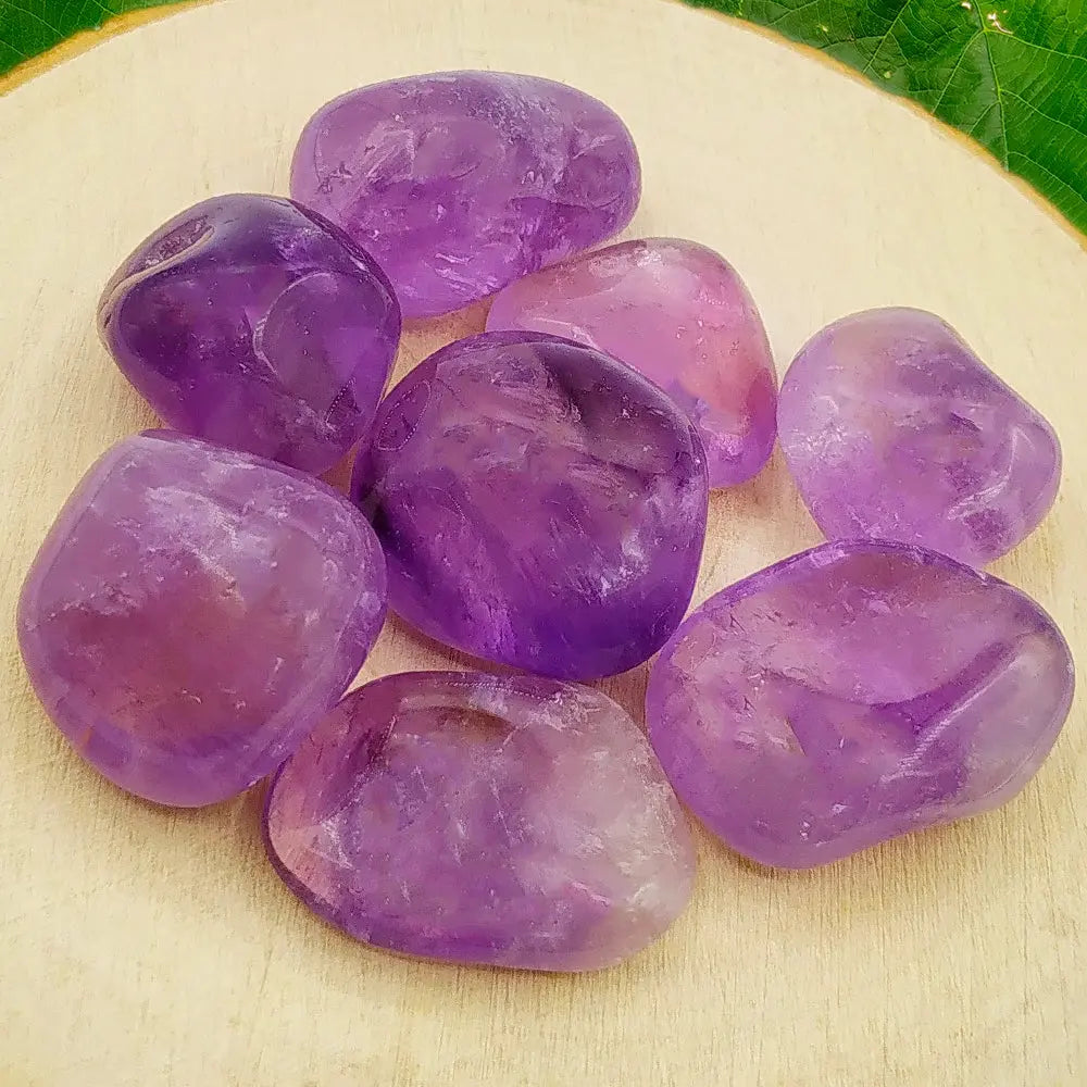 Un groupe de huit pierres d'améthyste roulées (taille M) aux teintes violettes variées et aux reflets cristallins, disposé sur un support en bois clair bordé d'une feuille verte.