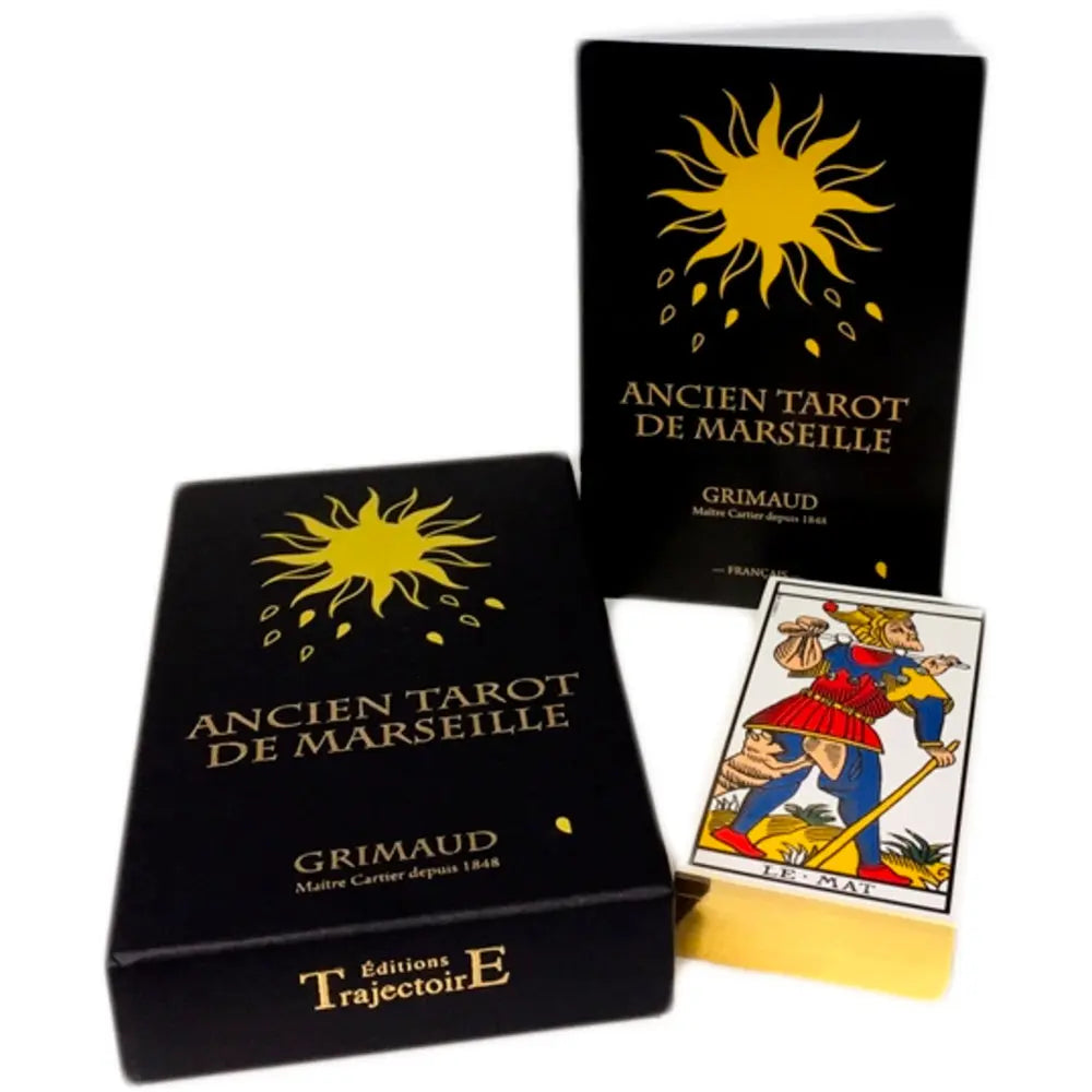 Coffret « Ancien Tarot de Marseille » (Grimaud) : deux boîtes noires avec un soleil doré et lettrage doré, dont une étui vertical « ANCIEN TAROT DE MARSEILLE » ; au premier plan, une carte du tarot « LE MAT » est partiellement sortie du coffret (Éditions TrajectoirE).