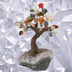 Arbre du bonheur décoratif avec un tronc en résine brune et des branches en fil doré ornées de pierres naturelles multicolores (cornaline, sodalite, aventurine...), fixé sur un socle en amas d'améthyste brute, devant un fond blanc aux motifs géométriques.