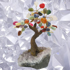 Arbre du bonheur aux feuilles en pierres naturelles multicolores et branches dorées, monté sur un socle d'améthyste brute, devant un fond blanc géométrique.
