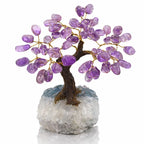 Un arbre du bonheur décoratif avec un tronc sombre texturé et des feuilles en améthyste violette montées sur fils dorés, reposant sur un socle massif de cristal de roche blanc.