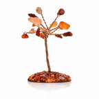 Un arbre du bonheur miniature composé d'un tronc en fils de cuivre torsadés et de feuilles en cornaline aux nuances orangées, reposant sur un socle couvert de petits éclats de pierres assorties sur un fond blanc avec reflet.