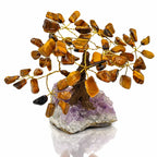Un arbre du bonheur décoratif aux feuilles en pierres d'œil de tigre et branches dorées, s'élevant d'un socle en amas de cristaux d'améthyste brute, présenté sur un fond blanc.
