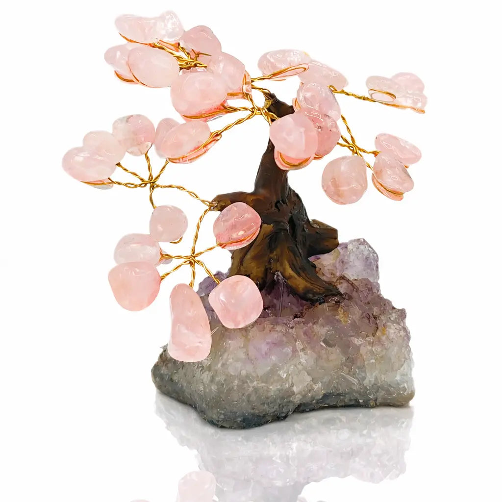 Un arbre du bonheur décoratif aux feuilles de quartz rose et branches dorées, s'élevant d'un socle en cristal d'améthyste brut, présenté sur un fond blanc.