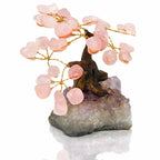 Un arbre du bonheur décoratif aux feuilles de quartz rose et branches dorées, s'élevant d'un socle en cristal d'améthyste brut, présenté sur un fond blanc.