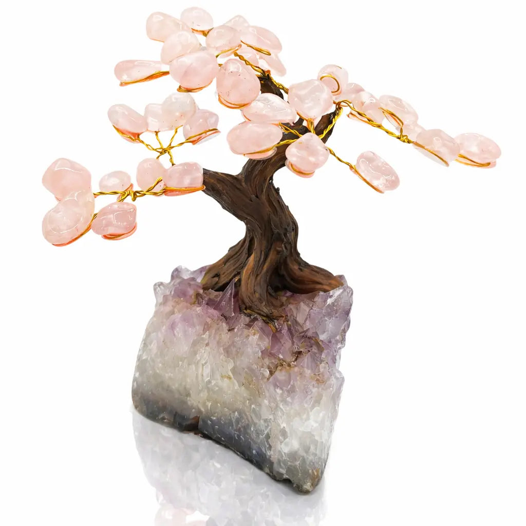 Un arbre du bonheur décoratif aux feuilles de quartz rose et tronc sculpté, s'élevant d'un socle en pointe d'améthyste brute, présenté sur un fond blanc.