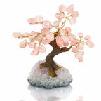 Un arbre du bonheur décoratif avec un tronc sculpté et des feuilles en pierres de quartz rose, solidement fixé sur une base en amas de cristaux de quartz, présenté sur un fond blanc.