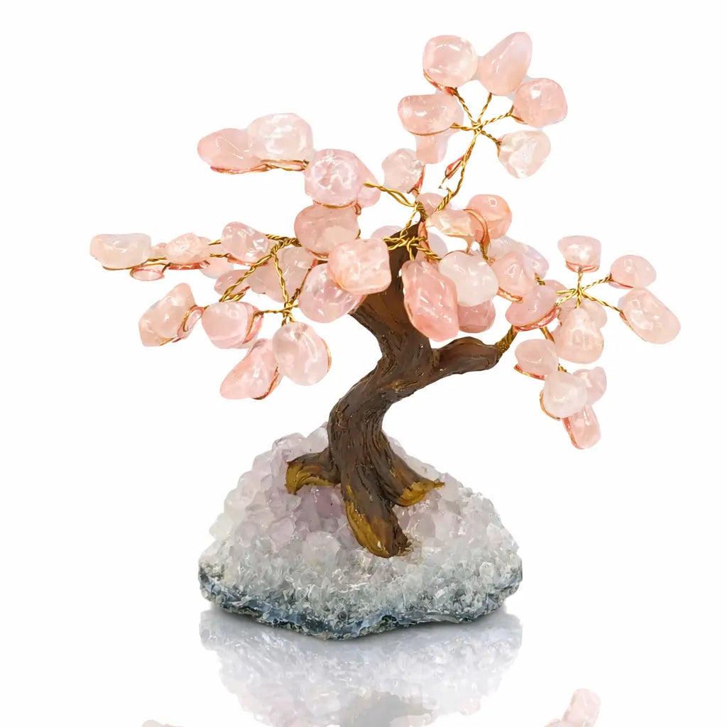 Un arbre du bonheur décoratif avec un tronc sculpté et des feuilles en pierres de quartz rose, solidement fixé sur une base en amas de cristaux de quartz, présenté sur un fond blanc.