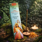 Boîte d'encens vert pâle Christ Miséricordieux Aromatika posée contre un tronc moussue en forêt, accompagnée d'un bâtonnet fumant, d'une bougie et de pierres d'ambre résineuses.