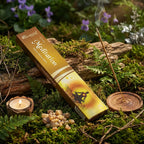 Boîte d'encens Méditation Aromatika illustrée d'une silhouette en lotus, posée dans un sous-bois moussu avec un bâtonnet fumant, une bougie et des grains de résine naturelle.