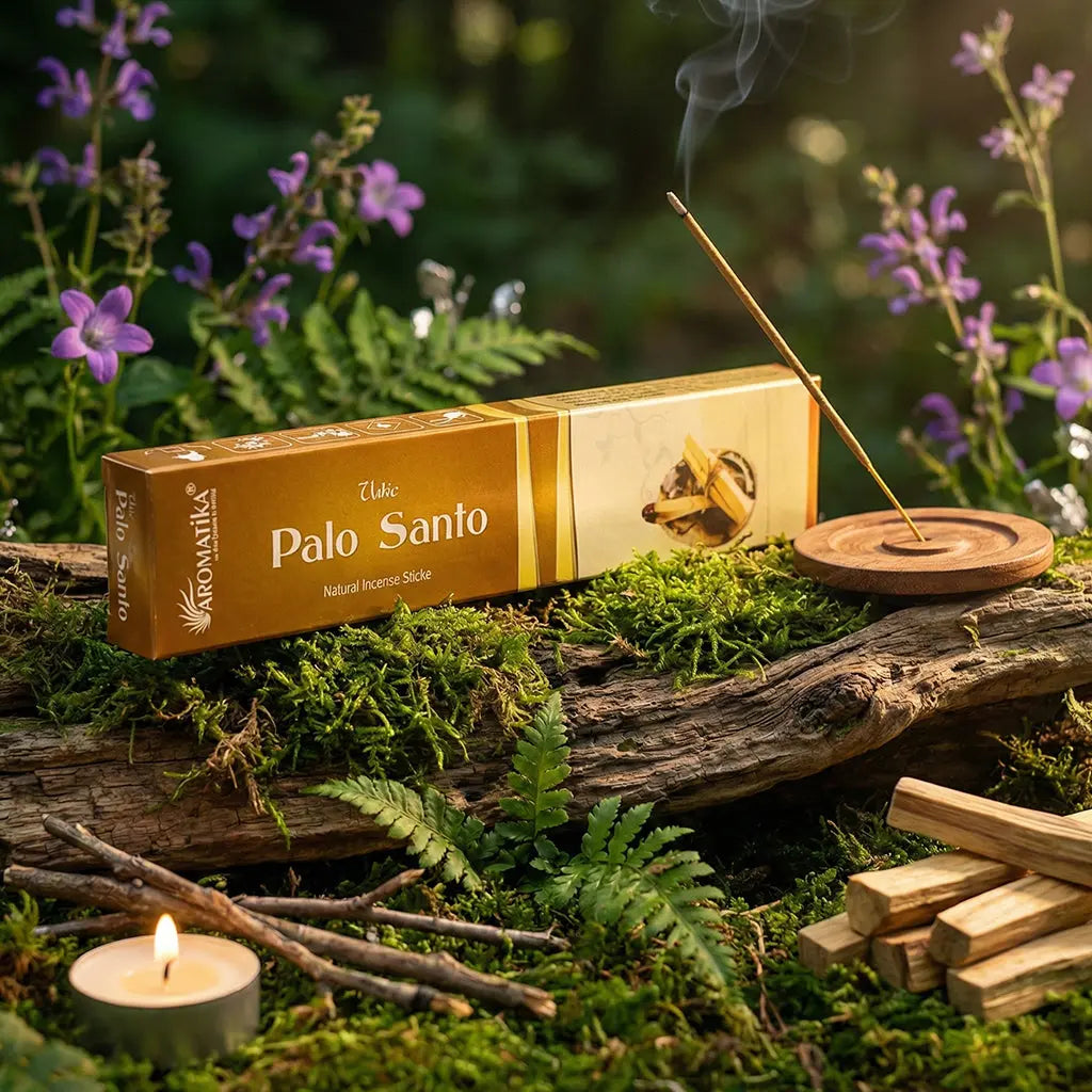 Boîte d'encens Palo Santo Aromatika déposée sur un lit de mousse verte et d'écorces, entourée de fleurs violettes et de petits rondins de bois sacré.