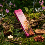 Boîte rose d'encens Saint Joseph Aromatika posée sur une branche moussue en forêt, entourée de fleurs violettes, d'une bougie et d'un tas de copeaux de santal rouge.