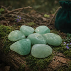 Ensemble de pierres d'aventurine verte polies d'un vert d'eau rafraîchissant, disposées sur un tronc d'arbre moussue avec un pochon vert sapin en arrière-plan.
